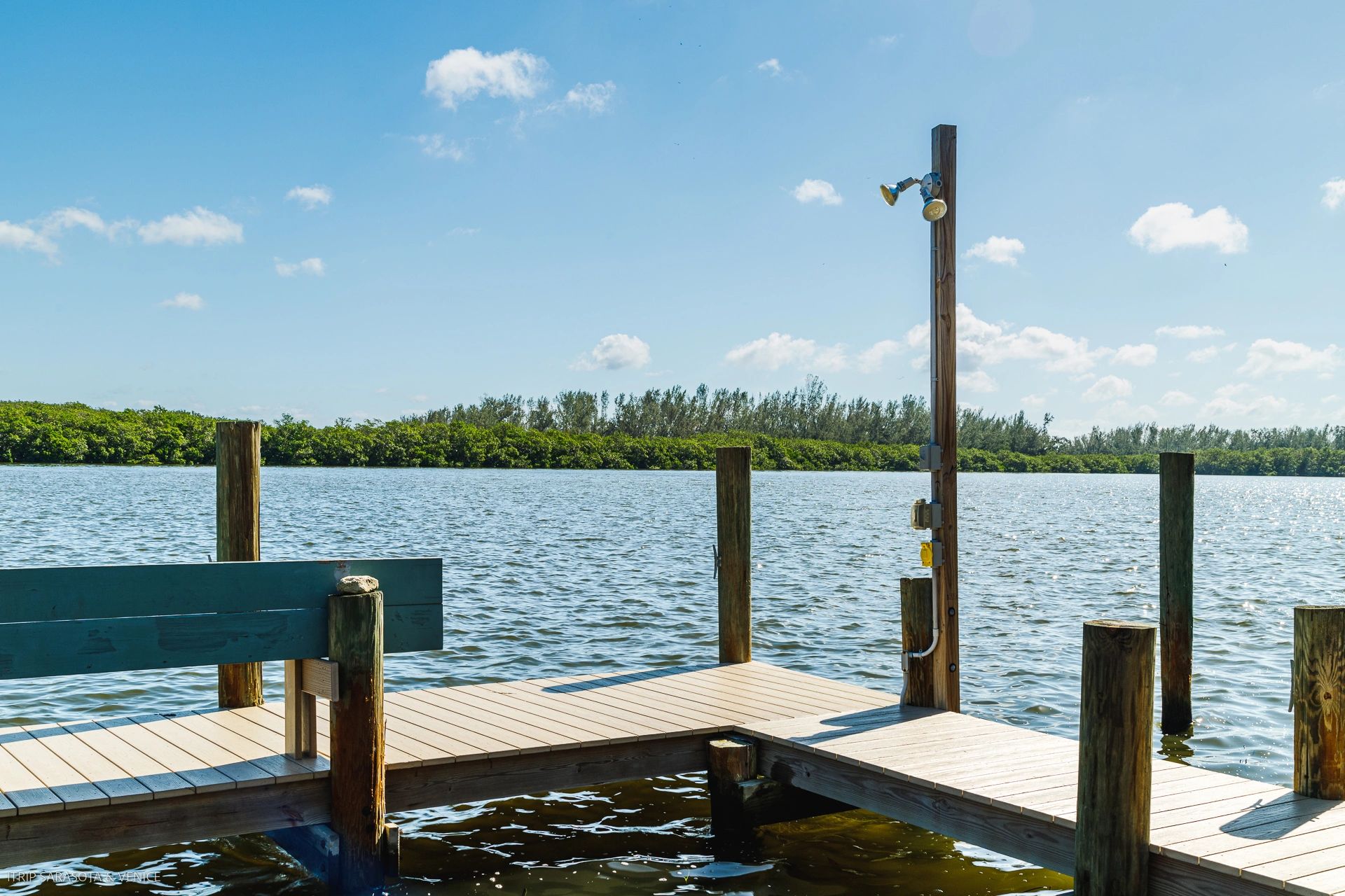 A wooden dock with an outdoor shower and bench overlooking a body of water with trees in the background.