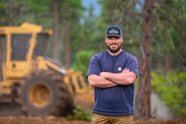 Man in navy shirt and cap standing with arms crossed near construction equipment.