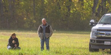 Two men in a field near a parked truck on a sunny day.