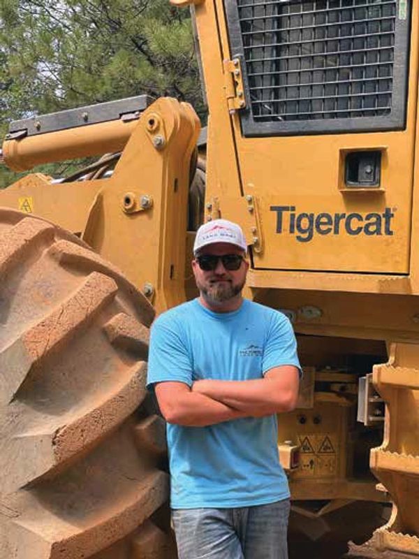 Man in blue shirt and cap stands with arms crossed by large Tigercat machine.