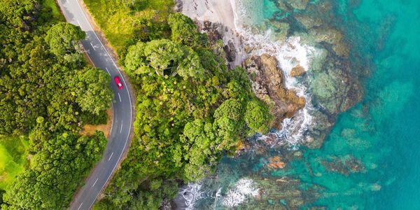 Aerial view of a winding coastal road beside blue‑green Mediterranean shoreline with lush trees