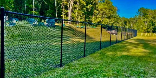 A long black chain-link fence borders a grassy area near a cemetery with trees in the background.