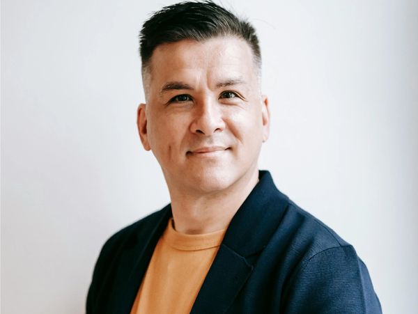 Man with short hair wearing a navy blazer and mustard shirt smiling.