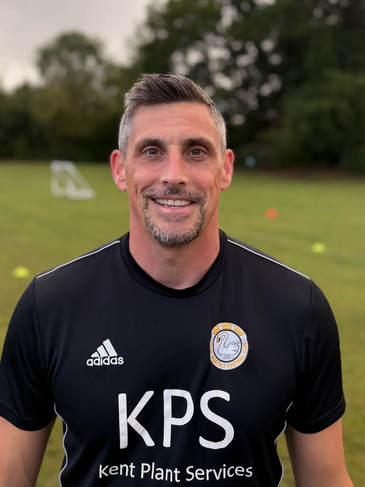 Smiling man in a black sports jersey on a grassy field with trees in the background.