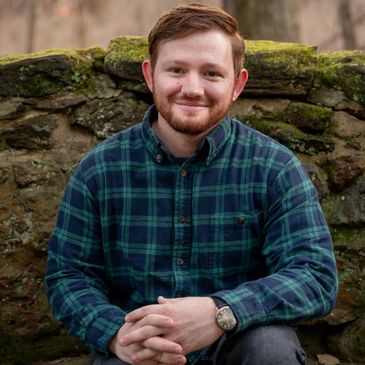 Smiling man in a green plaid shirt sitting outdoors against a mossy stone wall.