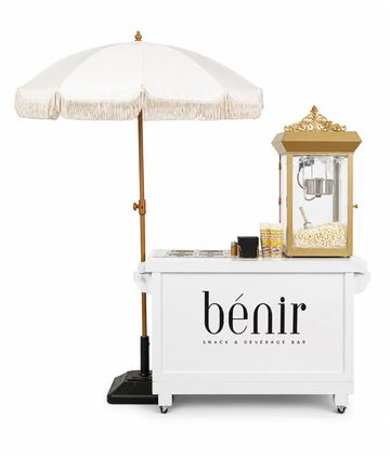 A white snack and beverage bar cart with a popcorn machine and umbrella.