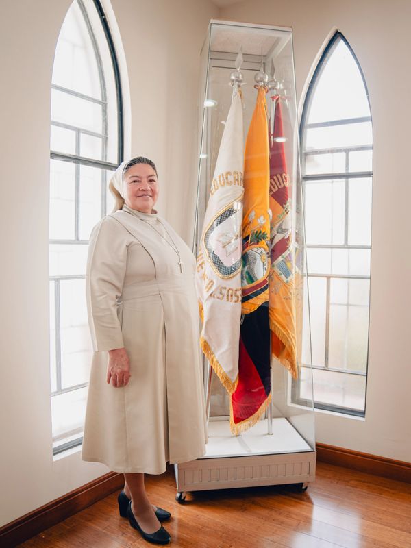 A nun in beige habit stands beside a glass case with flags in a bright room.