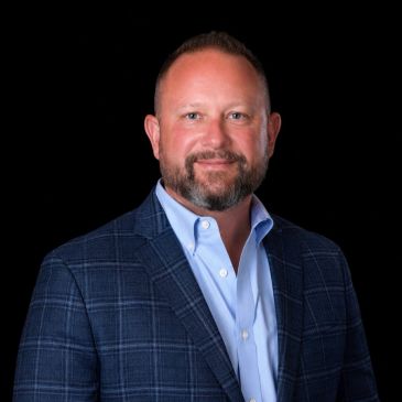 Man in a blue checkered blazer and light blue shirt against black background.