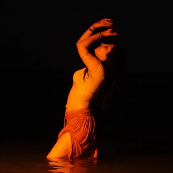 Woman in water illuminated by warm light, posing with arms raised against a dark background. Hippie, inspirational, spiritual, 