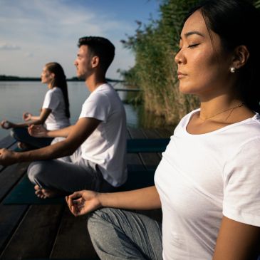 Three people meditating outdoors by the water.