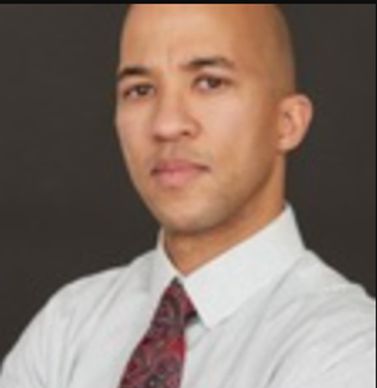 Dark-skinned man pictured behind blurred background, white shirt, red tie.