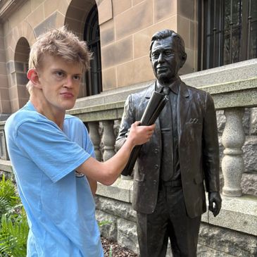 A young man in blue shirt playfully holding newspapers near a statue outside a building.