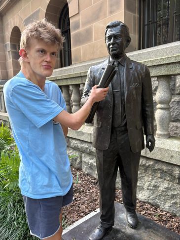 A young man in blue shirt playfully holding newspapers near a statue outside a building.