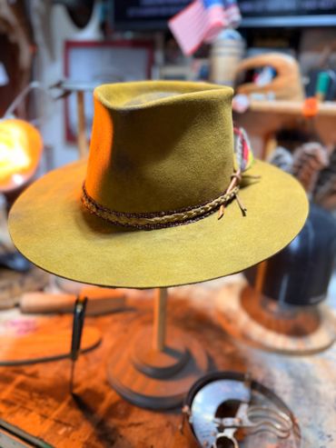 A stylish mustard felt hat with braided band and feather on a wooden stand.
