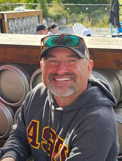 Man in sweatshirt with baseball cap and sunglasses smiling for the camera