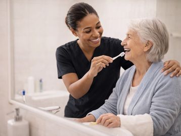 Caregiver helps elderly woman brush her teeth with a smile.
