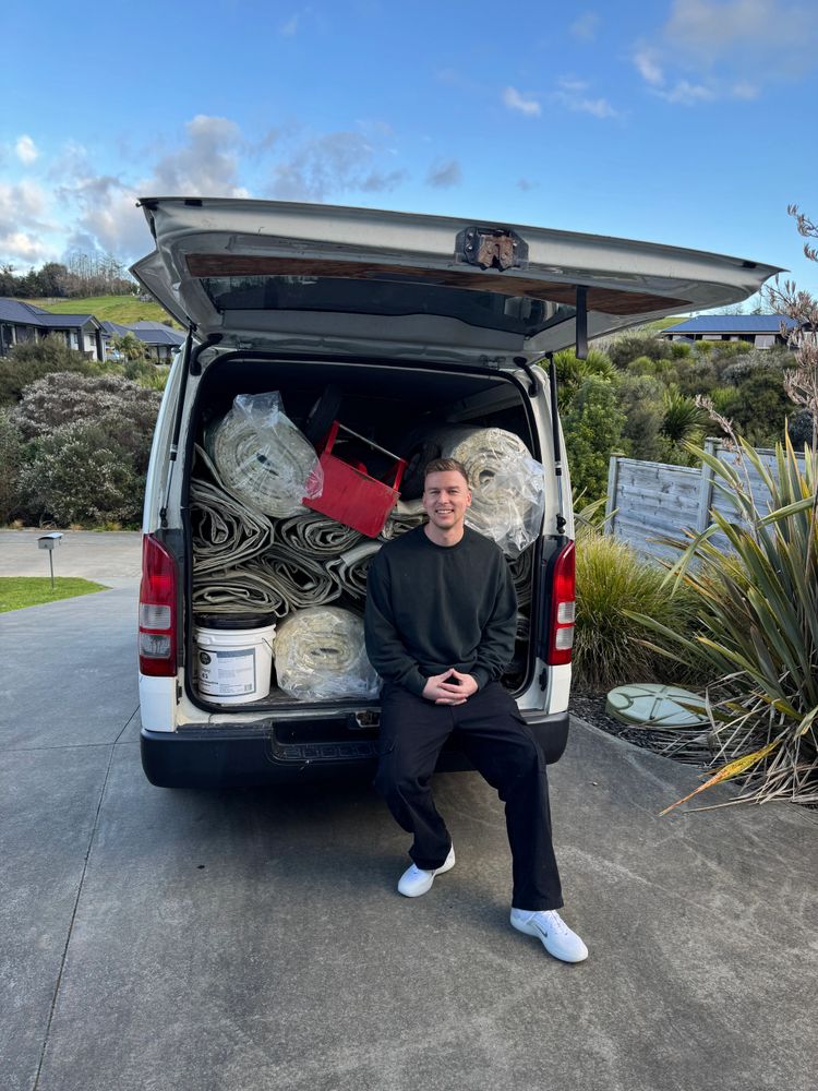 A man sitting on the back of a packed white van with rolled carpets and tools.