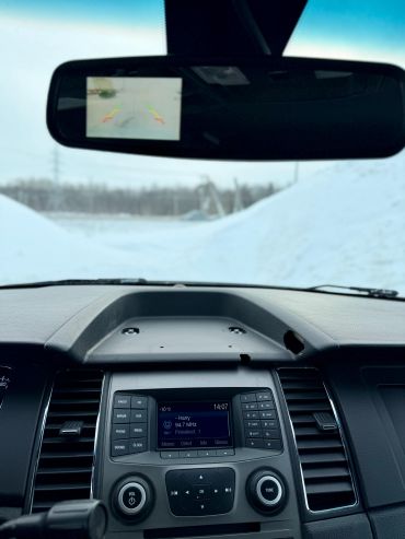 Car dashboard with rearview mirror showing backup camera in snowy environment.