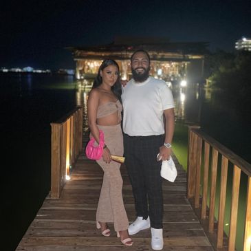 A couple posing on a wooden pier at night with lights in the background.