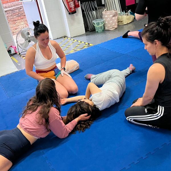 Four women on a blue mat practicing relaxation or first aid techniques indoors.