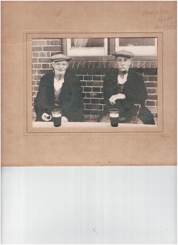 Two elderly men in flat caps sit outdoors with pints of beer, one smoking a pipe.
