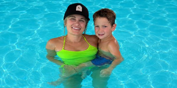 A woman in a neon green swimsuit and black cap holds a smiling boy in a blue swimsuit in a pool.