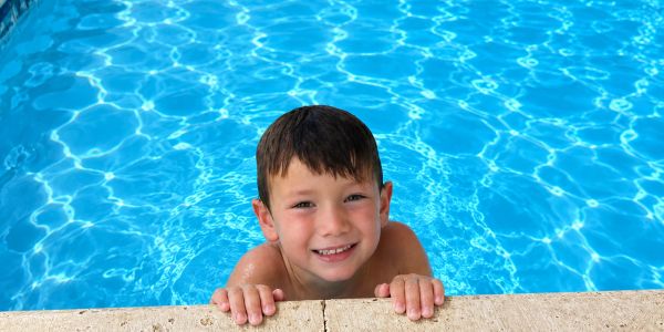 Happy boy smiling while holding pool edge in clear blue water.