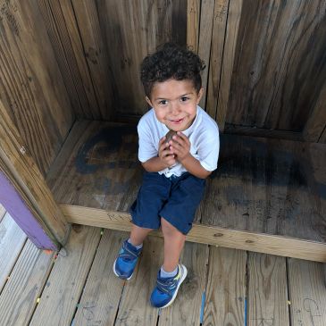 This image shows a young child wearing a white polo shirt and blue shorts while sitting on a wooden 