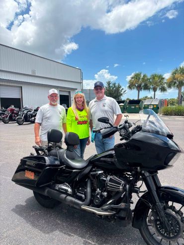 Three people standing behind a black motorcycle in a parking lot.