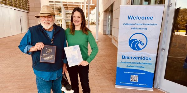 Two people standing near a California Coastal Commission public hearing welcome sign.