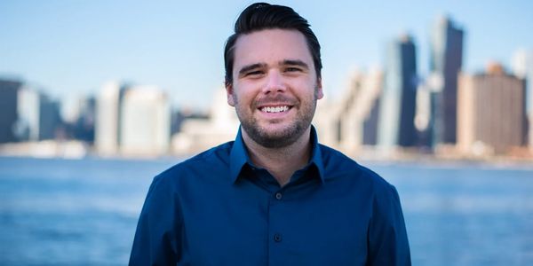 Smiling man in a blue shirt with city skyline and water in background.