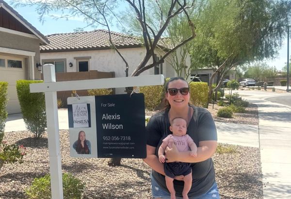 Woman holding baby next to real estate sale sign.