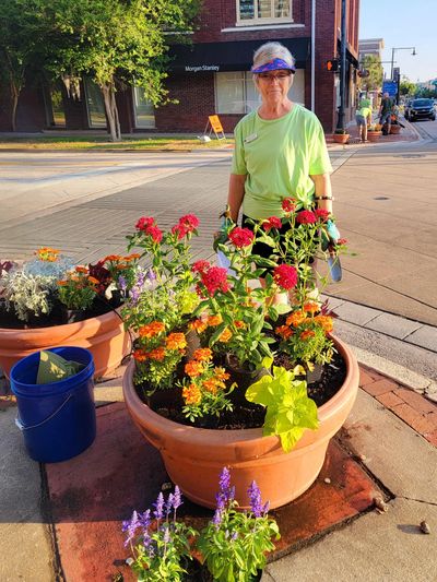 Woman standing behind large flower pots on a sunny street corner.