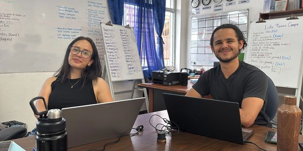 Two colleagues smiling while working on laptops in an office with whiteboards behind them.