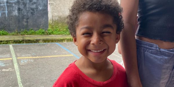 A smiling young boy in a red shirt stands outdoors next to someone.