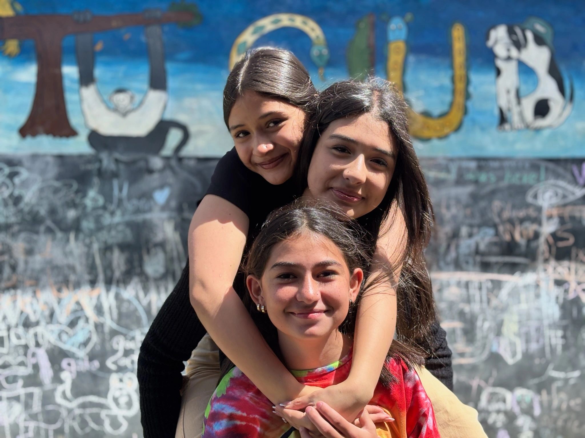 Three smiling girls hugging in front of a colorful graffiti wall.