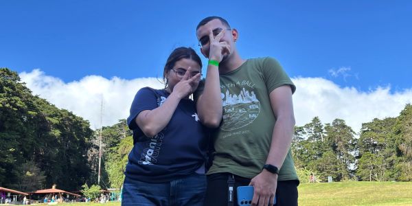 Two people posing outdoors with a bright blue sky and greenery.