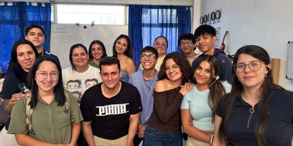 A group of smiling young adults posing indoors with blue curtains in the background.
