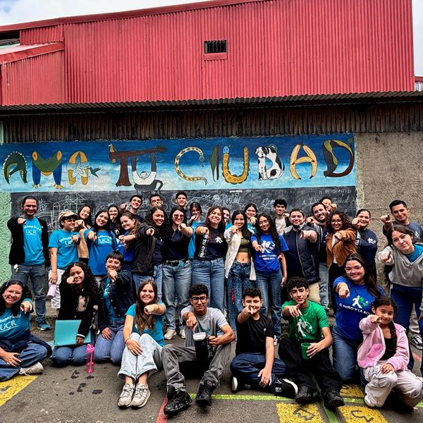 Group of diverse young adults posing in front of a colorful mural saying 'Ami Tu Ciudad'.