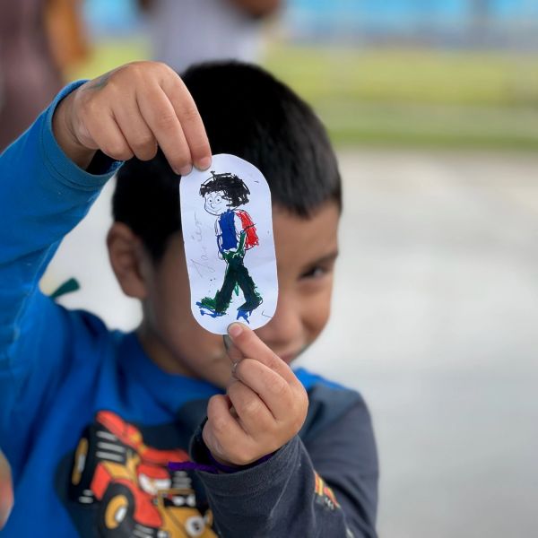 A child proudly holds up a hand-colored drawing of a person with a backpack.