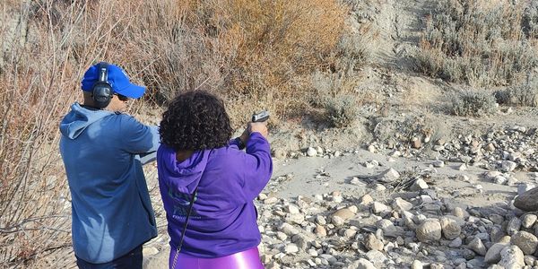 Two people aiming handguns outdoors in a rocky area.