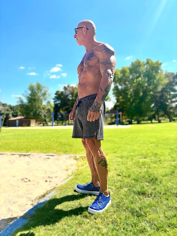 Tattooed muscular man standing outdoors in blue shoes and sunglasses on a sunny day.