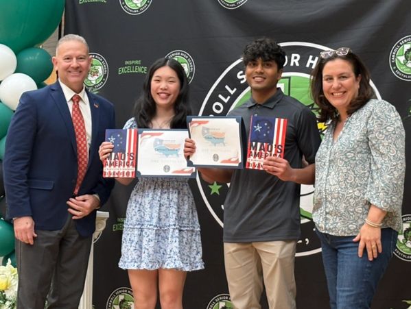 Two students hold certificates and awards, flanked by two adults at a school event.