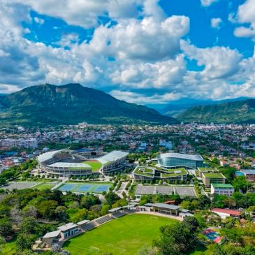 A stadium and buildings with mountains and a city under a cloudy sky.