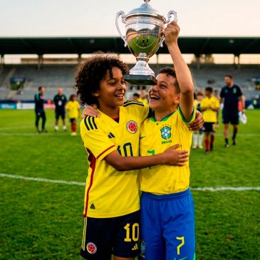 Two young soccer players from Colombia and Brazil celebrate with a trophy on the field.