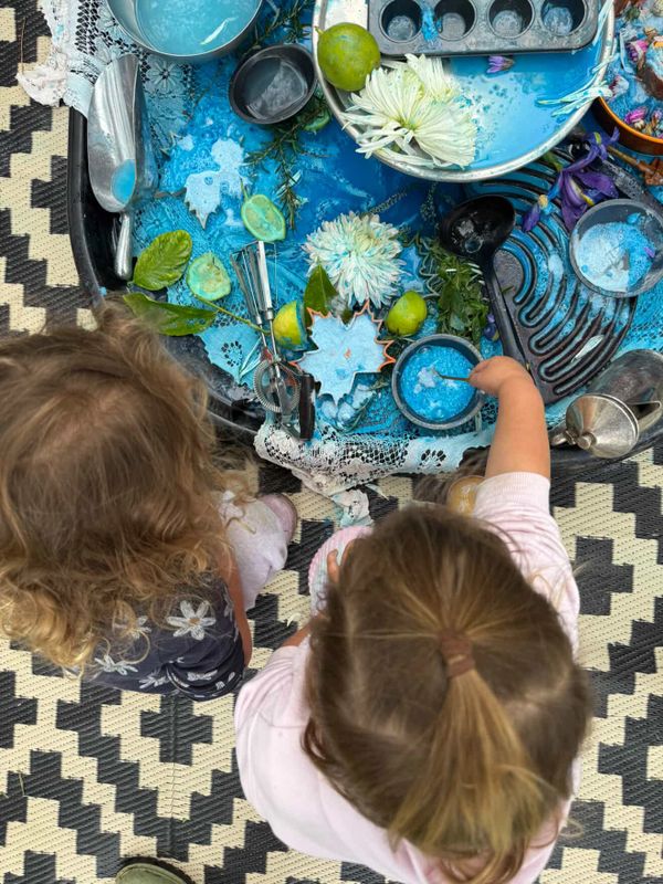 Two children explore a sensory tray filled with blue paint, leaves, and kitchen utensils.