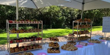 Outdoor catering setup with sandwiches, pastries, and fruits under large white umbrellas.