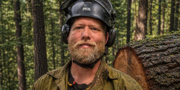Portrait of founder Chris Oliver in logging PPE standing in front of a cut down and stacked tree. 
