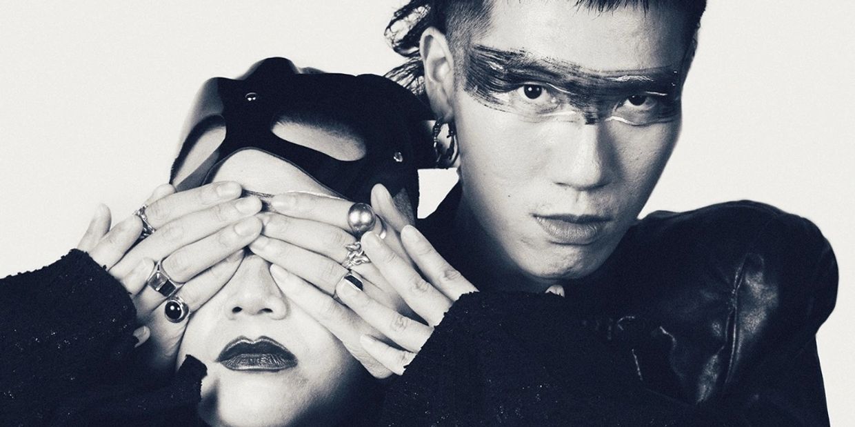 Two individuals in dramatic black and white fashion, one covering the other's eyes.