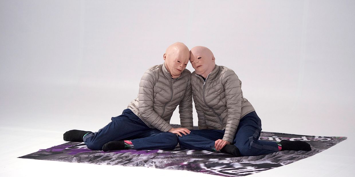 Two individuals in gray jackets and masks sitting closely on a patterned mat.